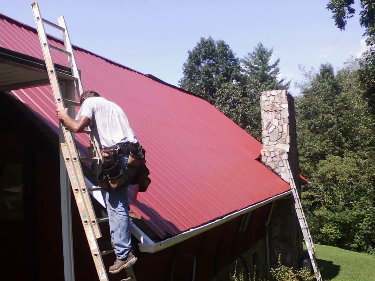 Roofer on a ladder beside a red metal roof and stone chimney — Soffit & Fascia Repair by Roofing Brothers in Shokan
